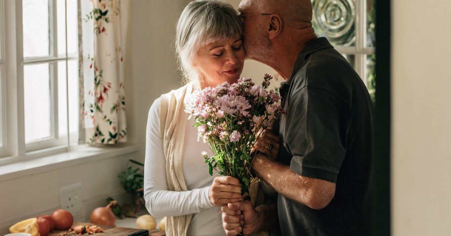 senior man kissing wife holding flowers by kitchen window